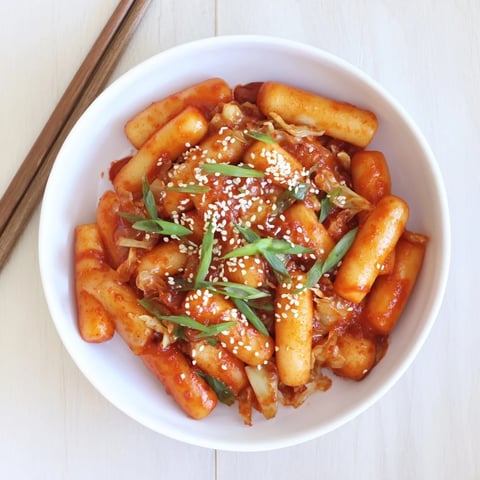 Steaming bowl of Korean Tteokbokki, featuring chewy rice cakes coated in a thick, vibrant red gochujang sauce, topped with fresh green onions and sesame seeds.  
