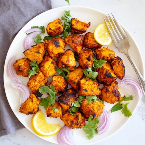 Golden-brown Indian Tikka Spiced Chicken pieces roasted on a wire rack, garnished with fresh cilantro and lemon wedges.  